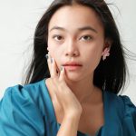 Close-up portrait of an Asian woman in a blue dress with pink flower earrings on a white background.